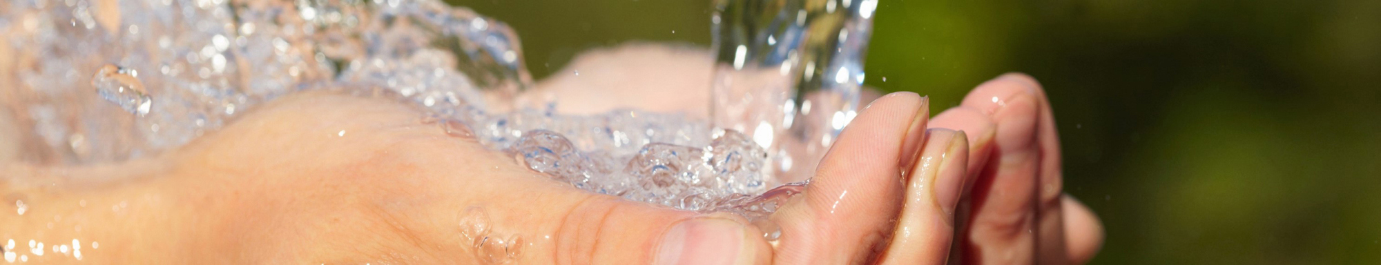 Hænder formet som en skål under rindende vand mod en sløret naturlig baggrund.
