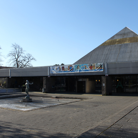 Moderne bygning med skråt tag, banner og statue foran, under klar himmel.