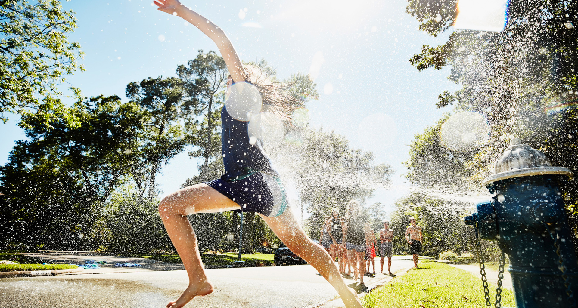 Barn løber glædesfyldt gennem vand fra en åben brandhane på en solrig dag.