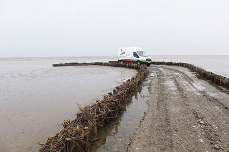Varevogn parkeret på en mudret vej nær havet, omgivet af sivbarrierer.