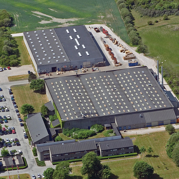 Luftfoto af en industrifacilitet med parkeringsplads og omkringliggende grønne områder.