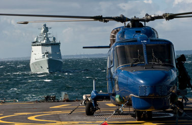 Blå helikopter på et skibs dæk med et flådeskib på vej.