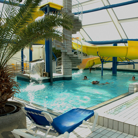 Indendørs pool med gul rutsjebane, palme og mennesker, der svømmer.