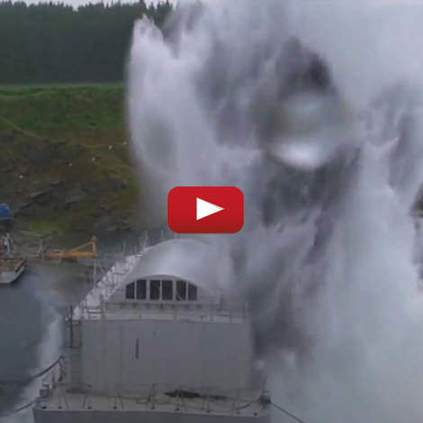 Stor eksplosion i vandet nær industriel struktur med skovklædt baggrund.