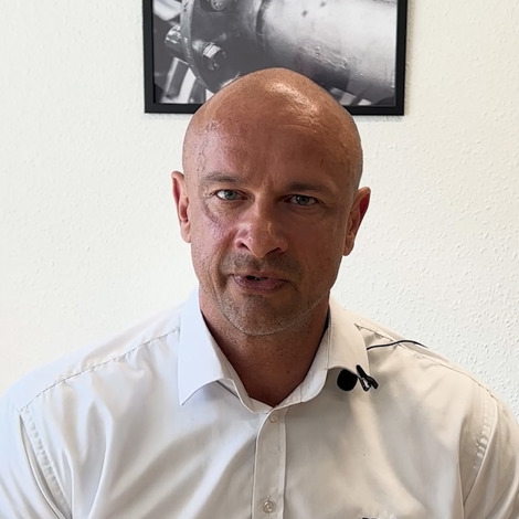 Mand i hvid skjorte taler, med industrielle fotos på væggen bag ham.