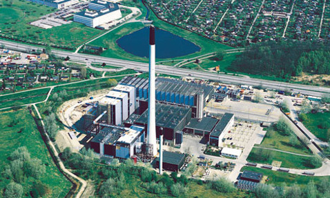 Luftfoto af et stort industrielt anlæg med en høj skorsten og omkringliggende grønne områder.