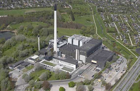 Luftfoto af en stor industrifacilitet med høje skorstene og omkringliggende grønne områder.