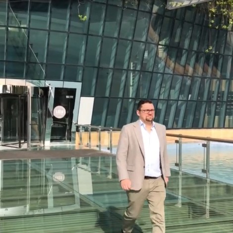 Mand går på en glasbro uden for en moderne glasbygning med grønne omgivelser.