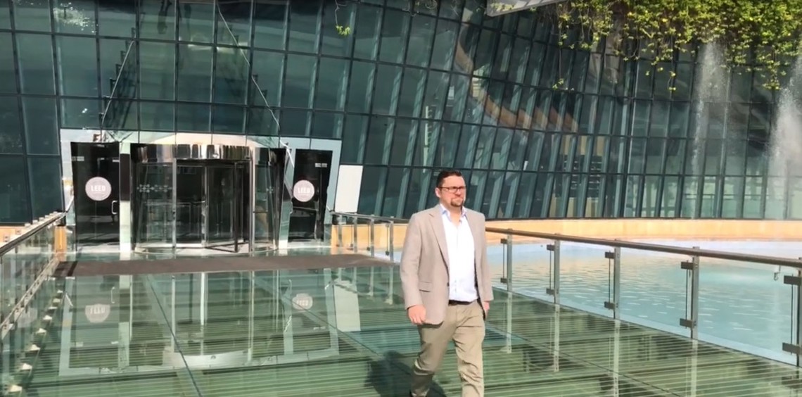 Mand går på en glasbro uden for en moderne glasbygning med grønne omgivelser.